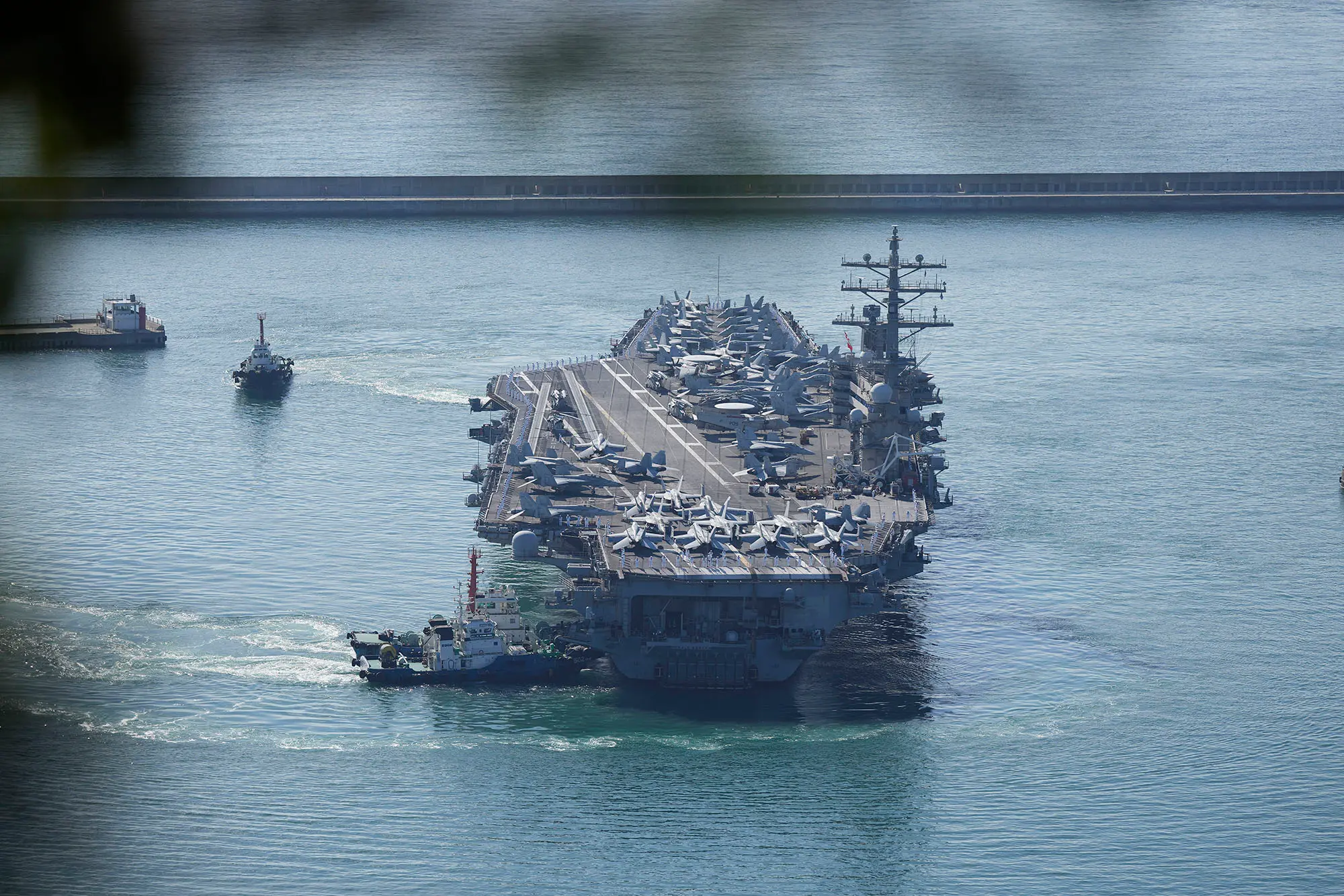 A wide aerial view of a calm blue harbor with a large gray vessel being guided into port, soft daylight and muted industrial tones.